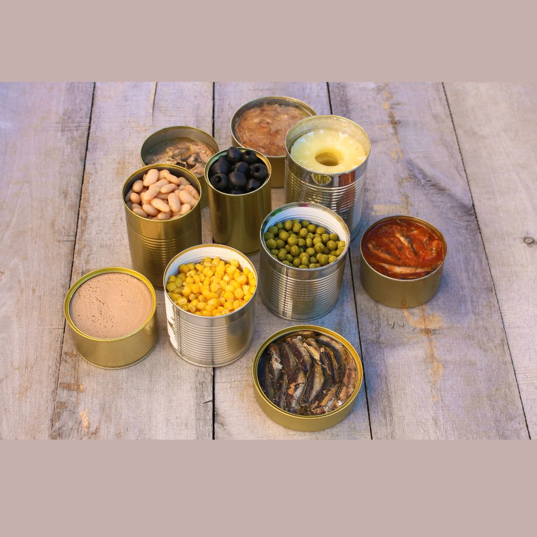L'image représente plusieurs boîtes de conserve ouvertes posées sur une table en bois. Elles contiennent divers aliments : des haricots blancs, des olives noires, du maïs, des petits pois, des tranches d'ananas, des sardines, et des champignons.