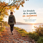 Une femme avec un sac à dos marche sur un sentier bordé d'arbres et de feuilles mortes au bord d'un lac, lors d'un coucher de soleil automnal.